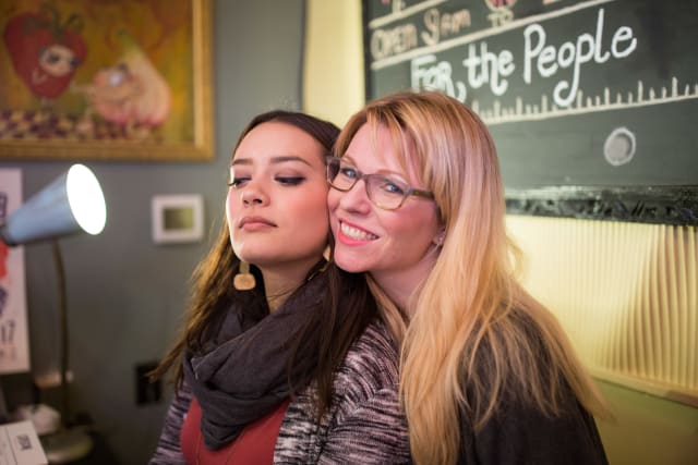 Two women posing together at the Love Bomb 2017 event, one with long dark hair and earrings, the other with blonde hair and glasses, against a backdrop featuring a chalkboard with the text "For the People."